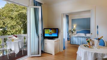 Ein Hotelzimmer mit Balkon, Fernseher, Bett mit blau-weiß gestreifter Bettwäsche, Esstisch mit Salat und einem Champagnerglas.