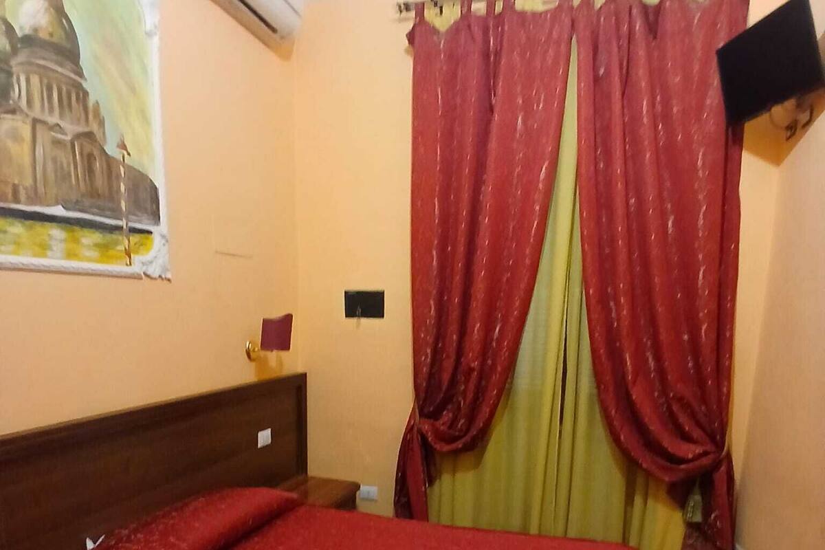 Une chambre d’hôtel avec un lit, un tableau représentant un bâtiment, un téléviseur accroché au mur et des rideaux rouges.