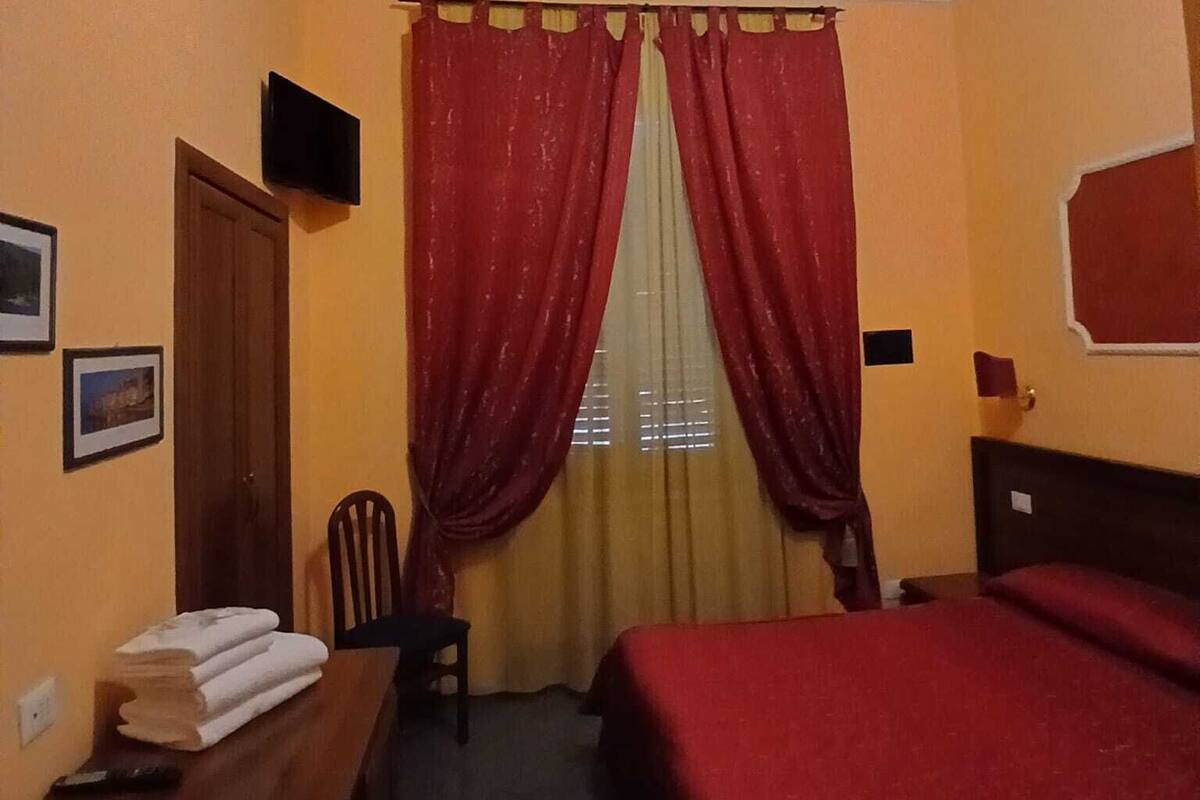 Une chambre d’hôtel avec un lit, un bureau et une chaise, un ventilateur de plafond et une fenêtre avec des rideaux.