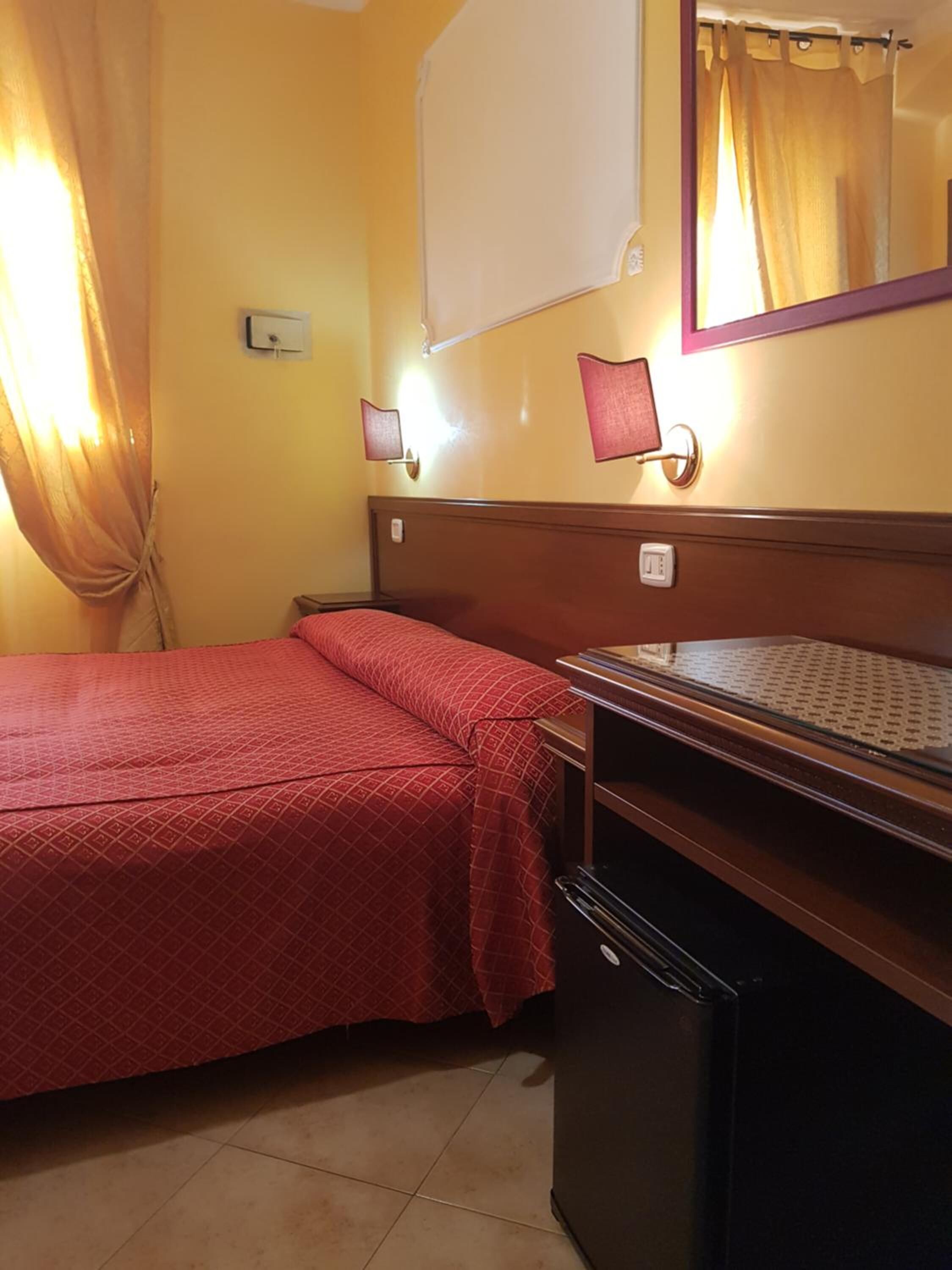 Une chambre d’hôtel avec un lit, un bureau, un miroir et une fenêtre avec des rideaux.