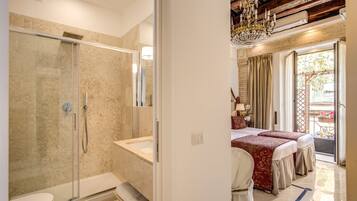 A hotel bathroom with a glass-enclosed shower, a bathtub, and a toilet. There is a bed with a red bedspread and a window with curtains.