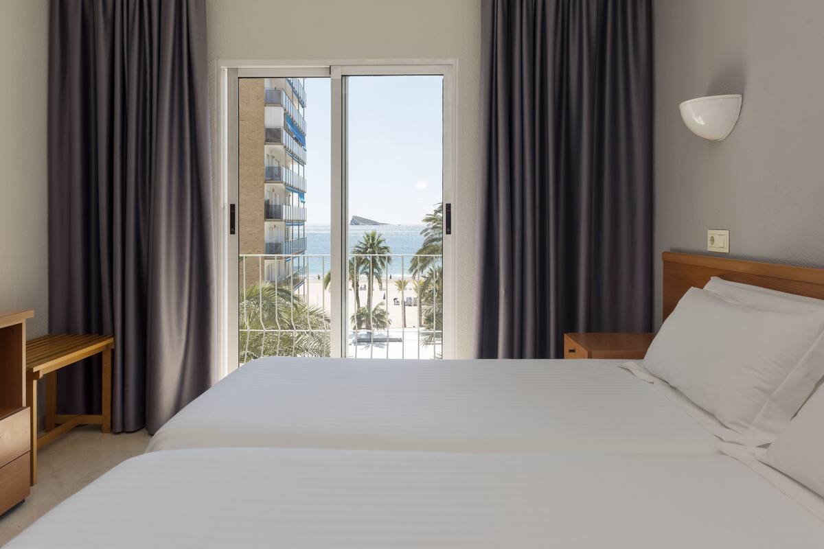 Quarto de hotel com cama, mesa de cabeceira de madeira, escrivaninha e vista para o oceano e palmeiras através de uma porta de correr.