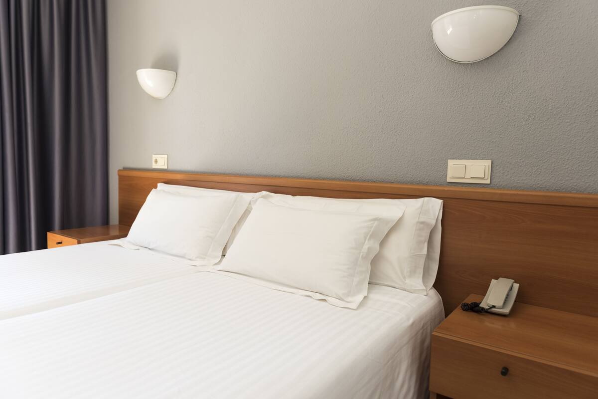 Quarto de hotel com uma cama bem arrumada, mesa de cabeceira com telefone e luminária fixada na parede.