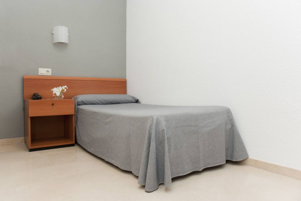 Uma cama de solteiro com colcha cinza em quarto com mesa de cabeceira de madeira, parede branca e abajur branco pequeno.