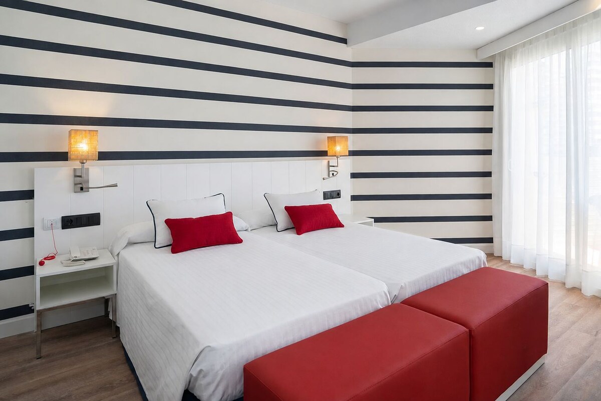 Habitación de hotel con dos camas, una mesita de noche con lámpara, un banco rojo y una ventana con cortinas.