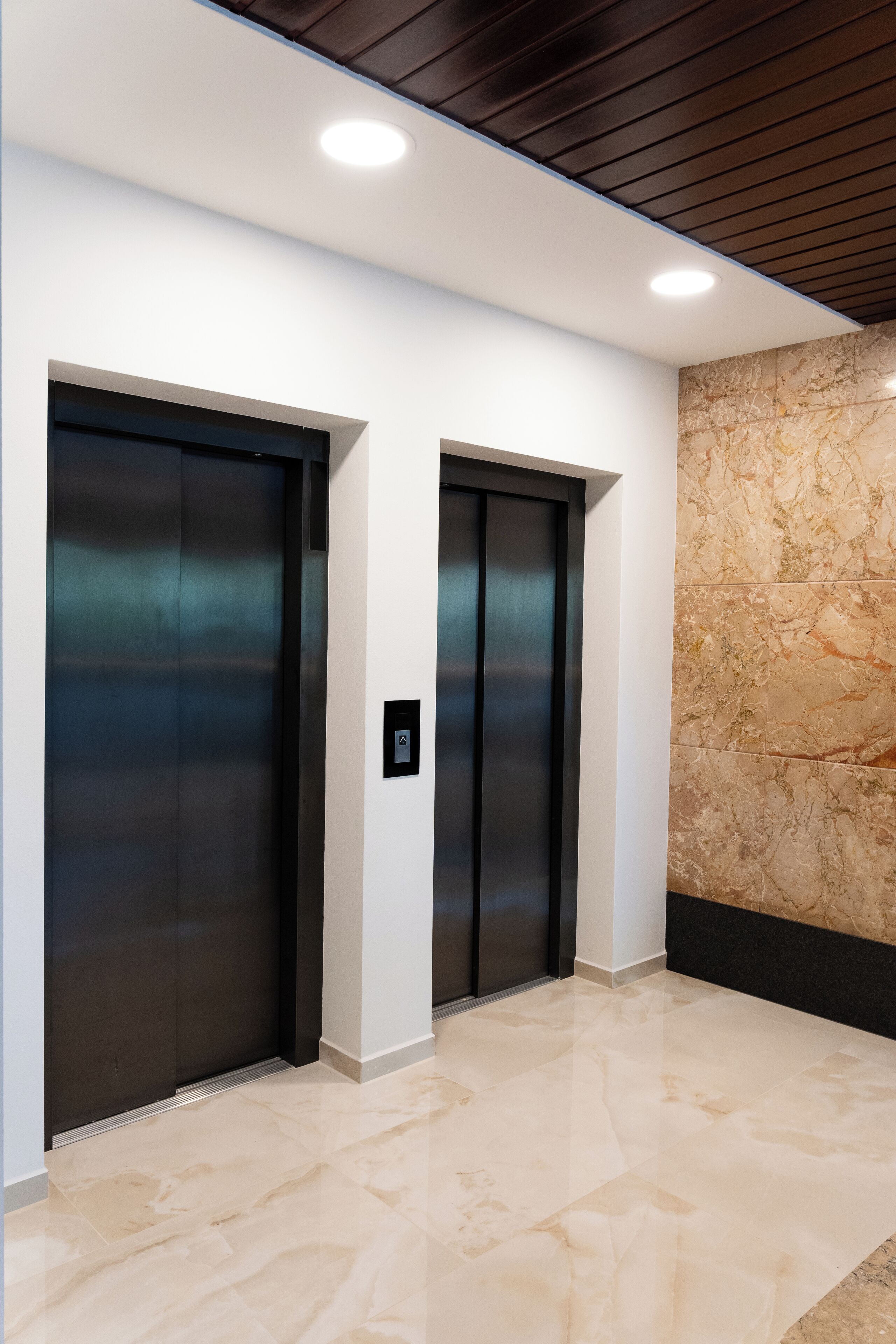 Two elevators in a hallway with a marble wall and a polished floor.