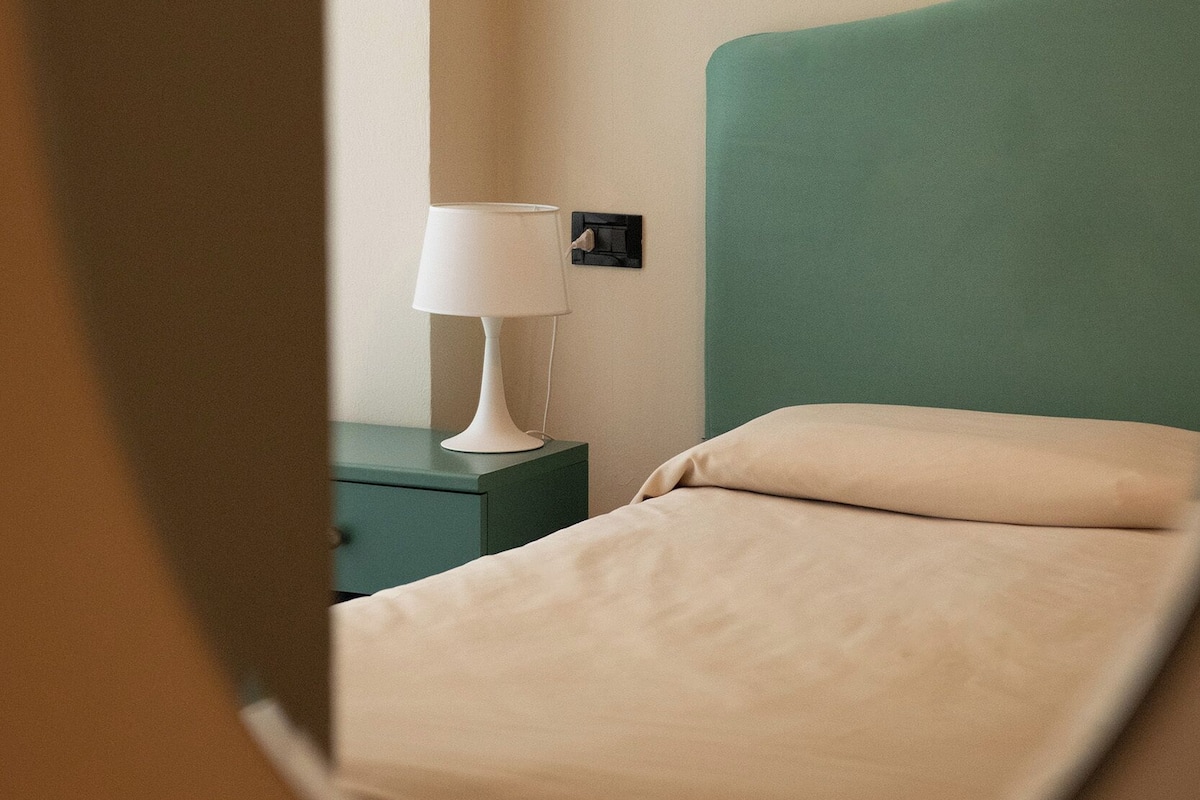 Una cama con cabecera verde y una lámpara de noche blanca en una habitación con pared color beige.