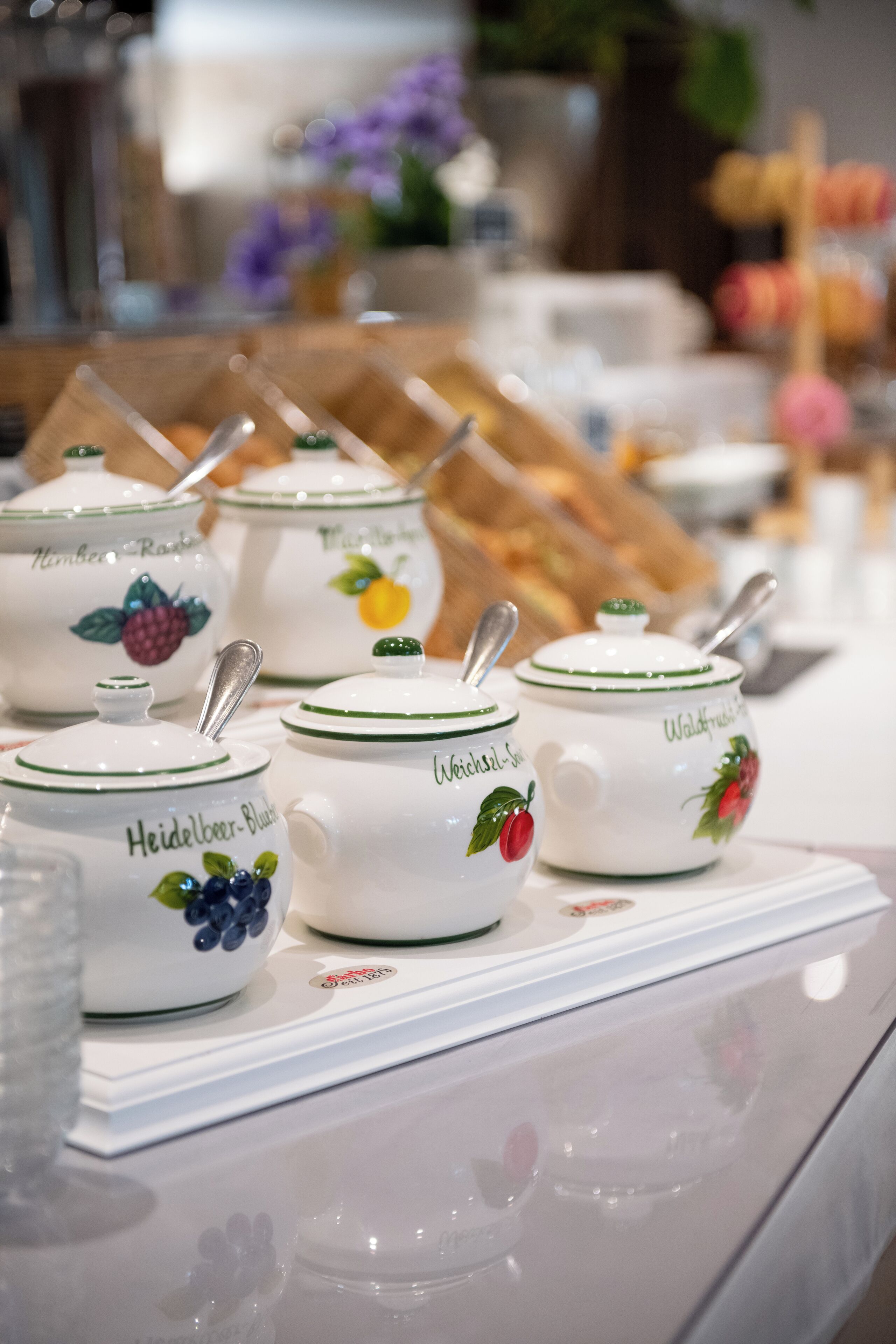 A display of white ceramic containers with green trim and fruit designs, labeled with names like "Heidelberger Blaue" and "Weichsel-Schnitzel."