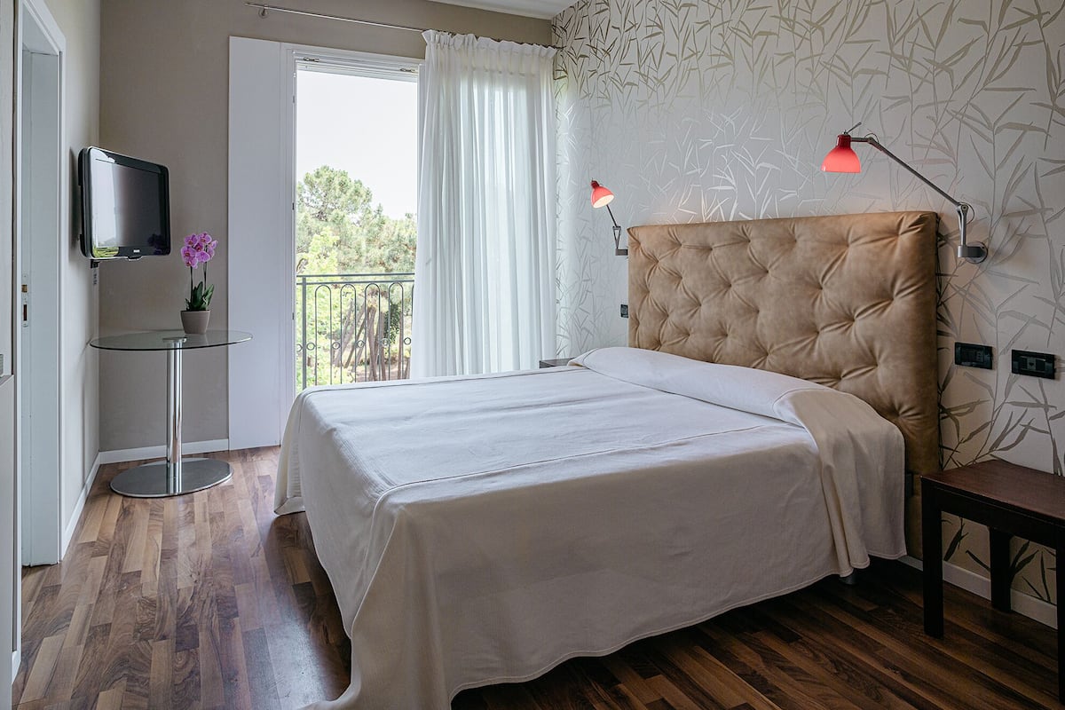 Un dormitorio con cama, televisor sobre un mueble bajo, una mesita con una planta y un balcón con cortinas.