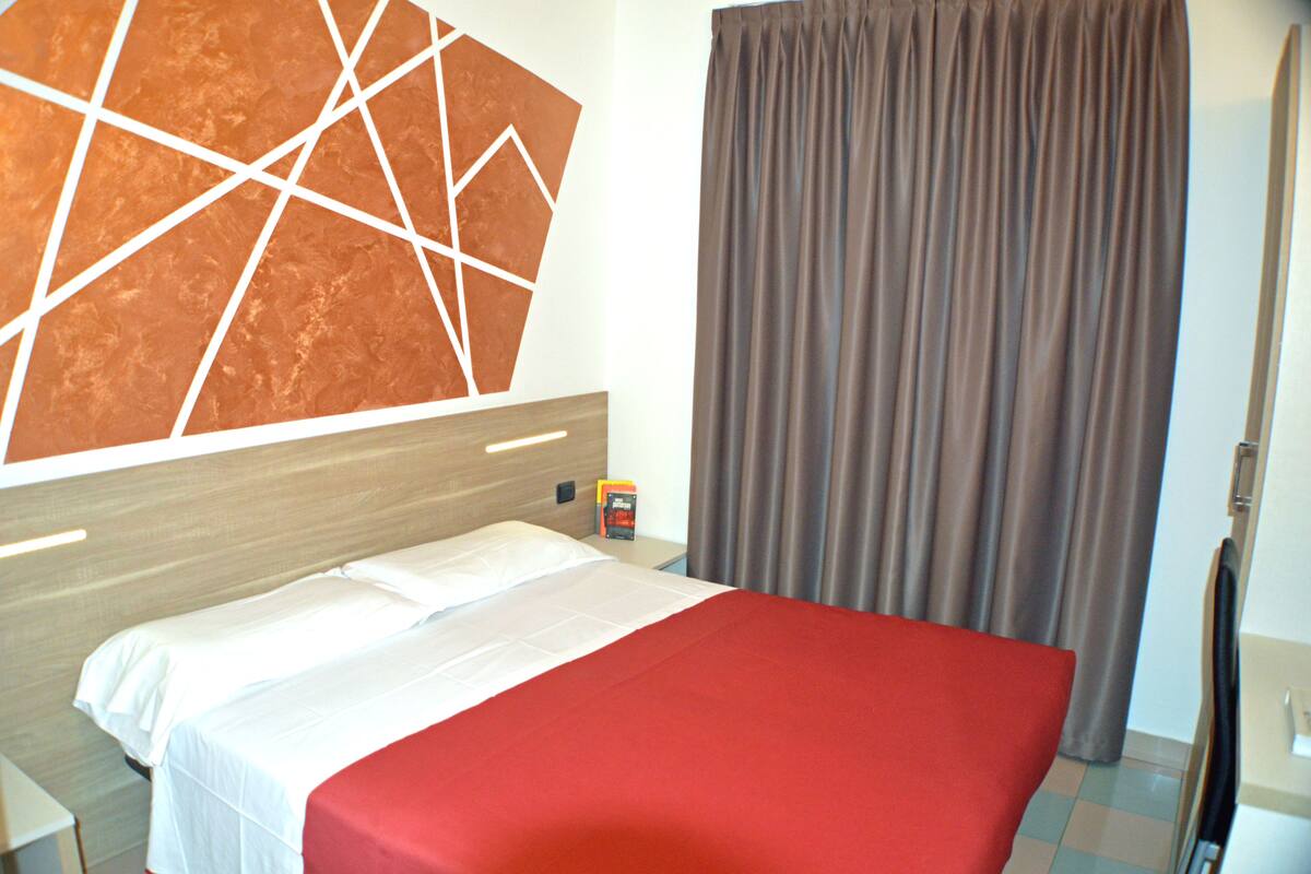 A hotel room with a bed, a wooden headboard, a red bedspread, a brown curtain, and a wall with a geometric design.