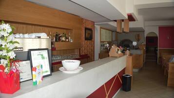 Zona de recepció d'hotel amb mostrador, diplomes emmarcades, un plat de fruita i un cistell de brossa.