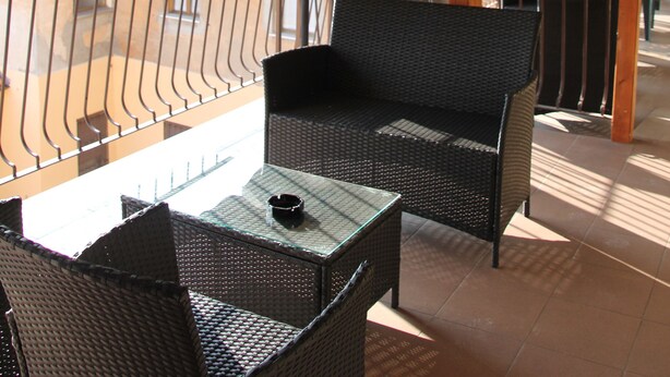 A covered outdoor seating area with wicker furniture and a glass table, featuring a view of a building in the background.