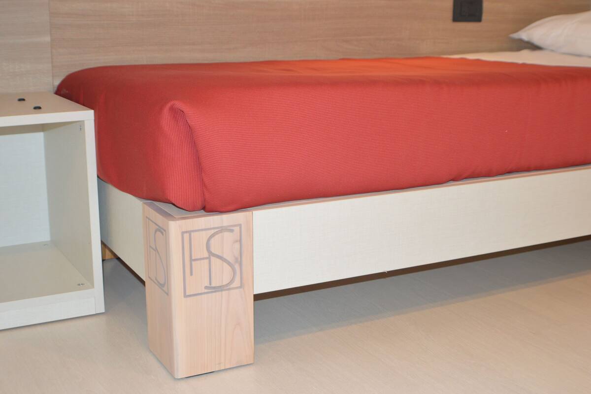 A bed with a red blanket, a white nightstand, and a wooden block with letters on it.
