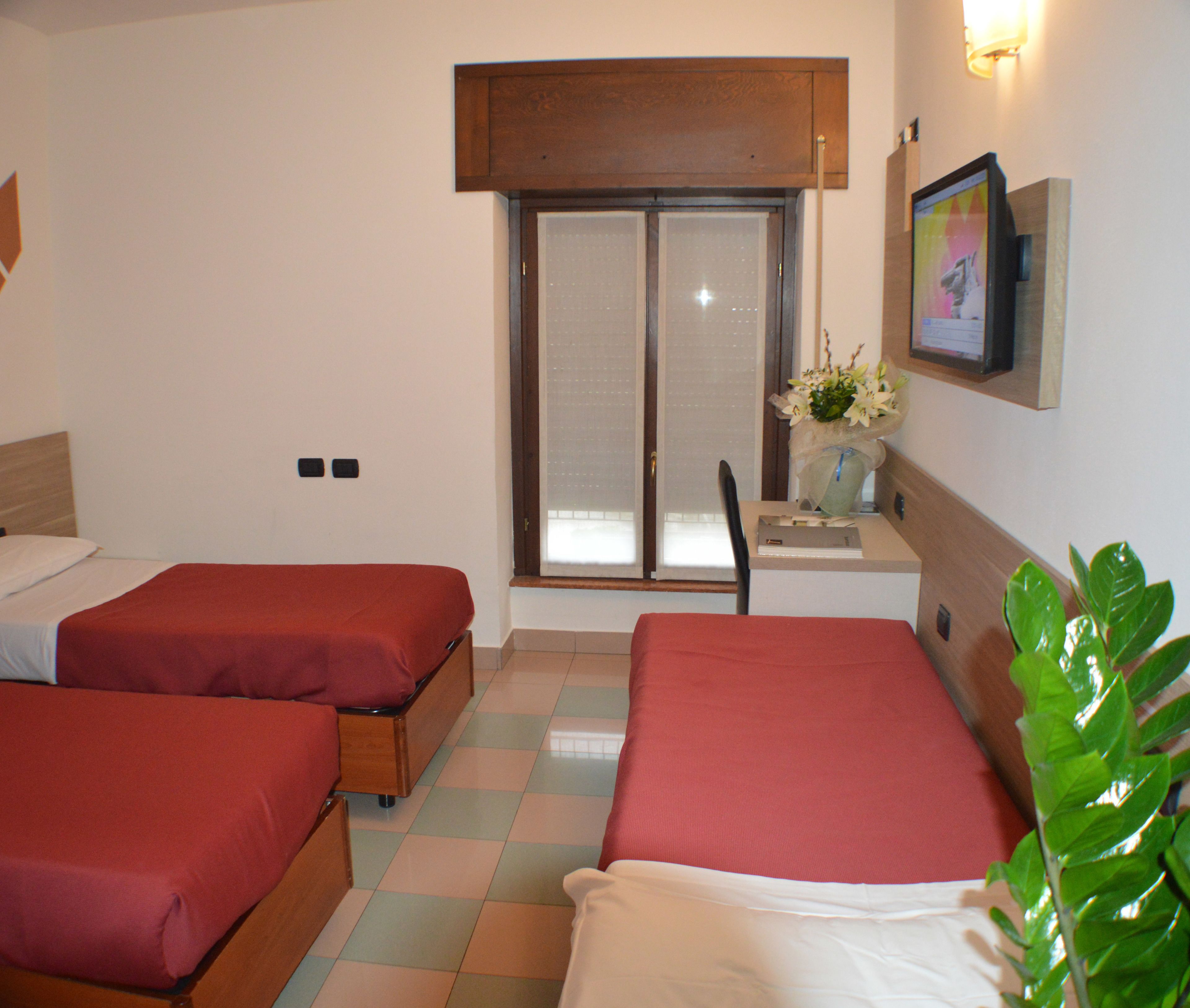 A hotel room with two beds, a desk with a chair, a window with blinds, a wall-mounted picture, and a potted plant.