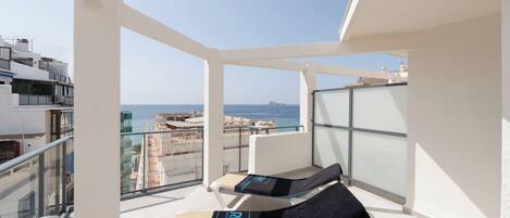 A balcony with two sun loungers, a glass railing, and a view of the sea and buildings.
