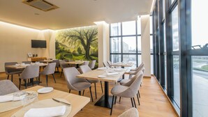 Un comedor moderno con varias mesas y sillas, un amplio ventanal con vistas, y un mural de un árbol en la pared.
