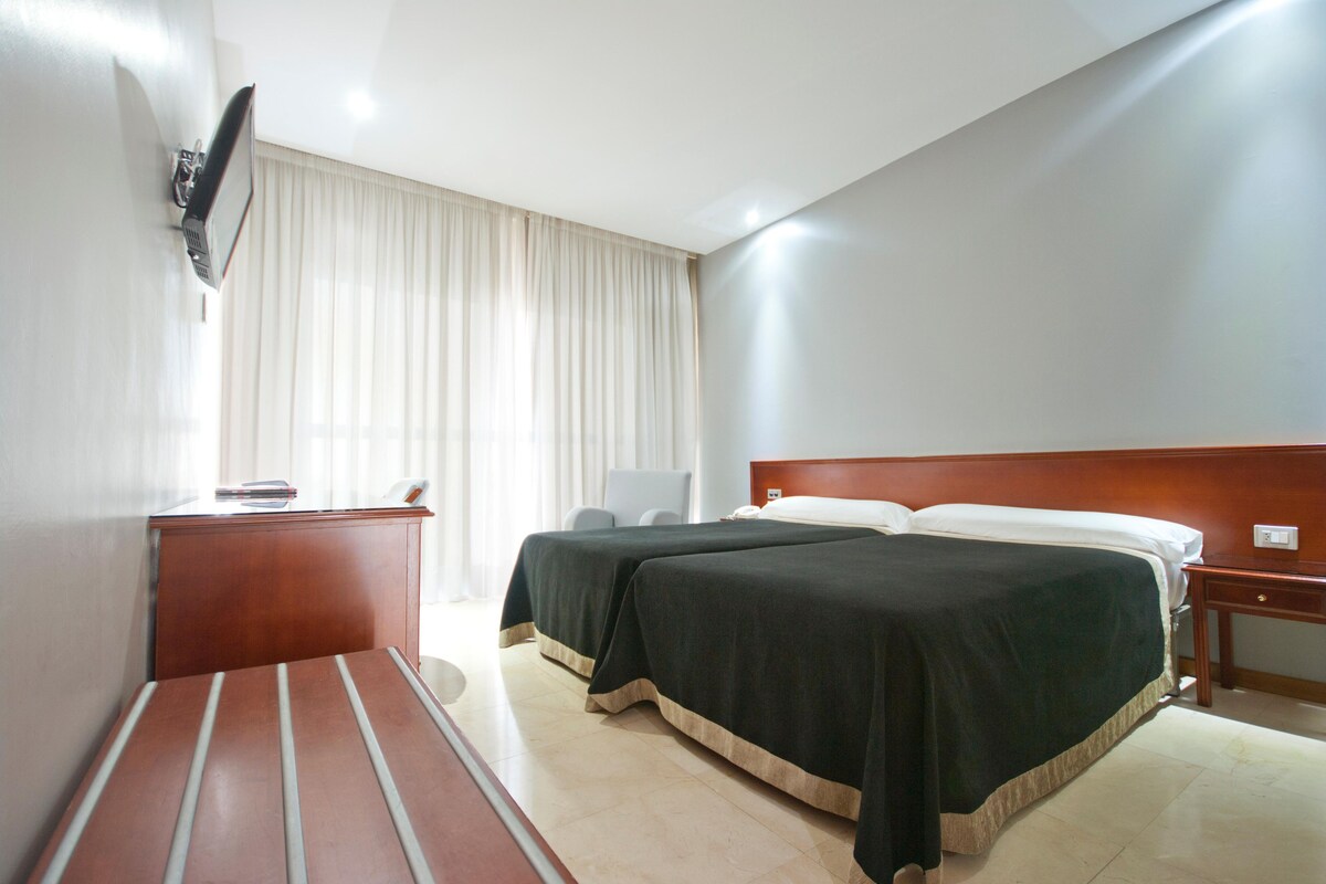 A hotel room with a bed, a desk, a chair, a television mounted on the wall, and a window with curtains.