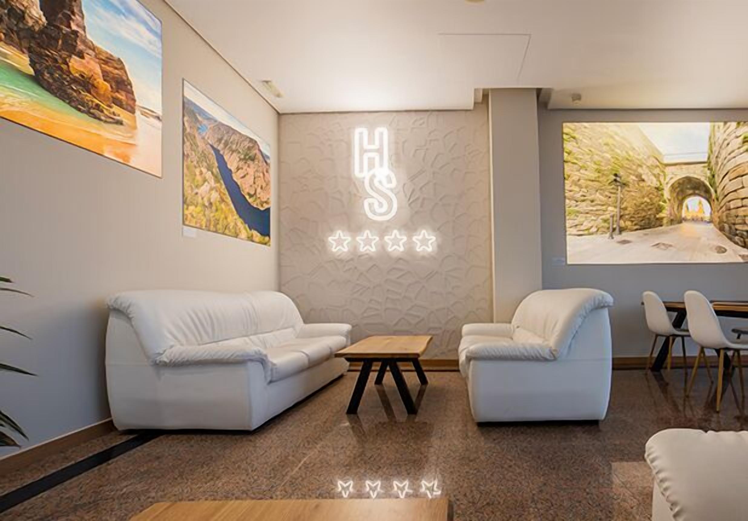 Un moderno vestíbulo de hotel con dos sofás blancos, una mesa de centro de madera y un aplique de pared que deletrea "HS".