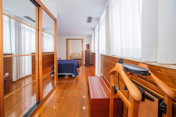 A hotel room with wooden flooring, a bed with a blue bedspread, a bench, a wooden desk, and a large mirror.