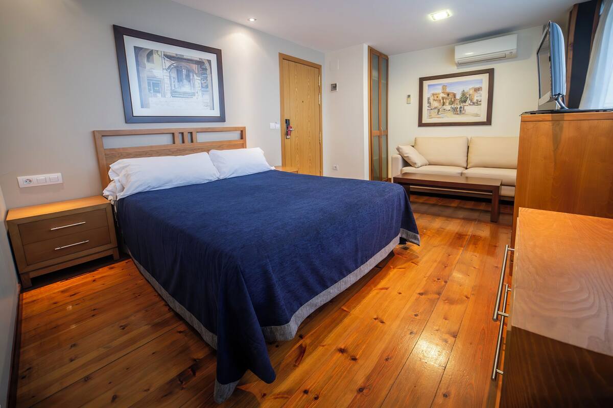 Een slaapkamer met een bed, houten vloer, een nachtkastje, een dressoir, een bank en lijstjes met foto's aan de wanden.