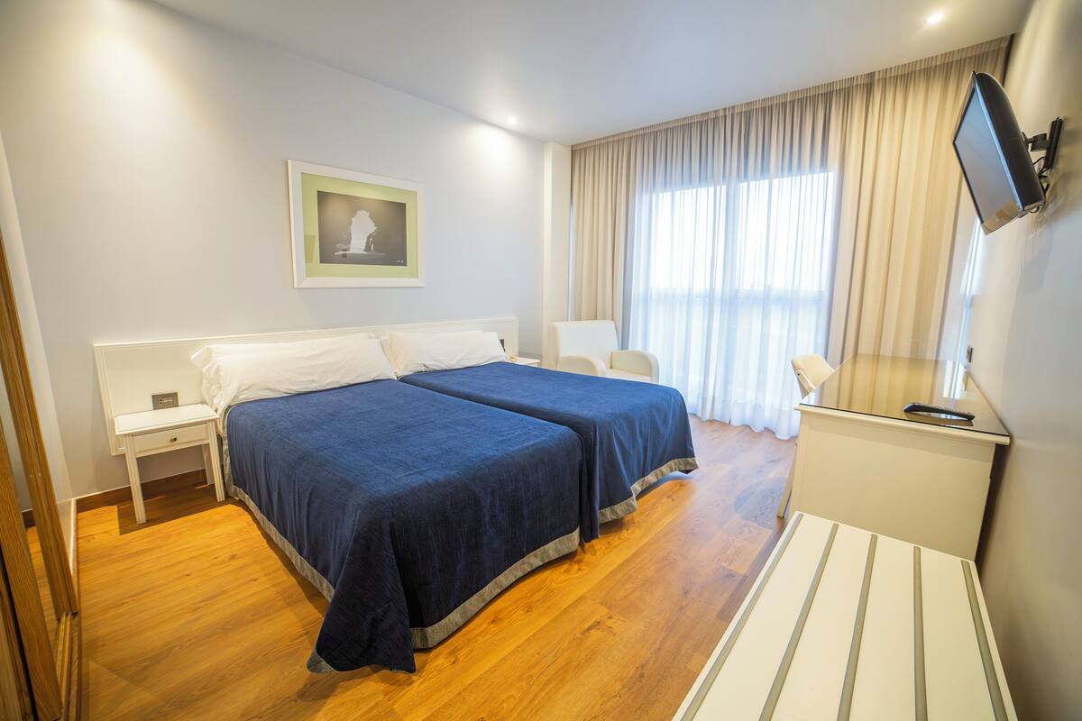 Hotelkamer met twee bedden, nachtkastje, stoel, televisie en raam met gordijnen.