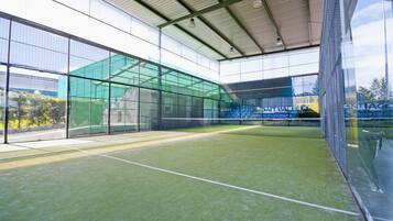An indoor tennis court with glass walls, a roof, and artificial turf.