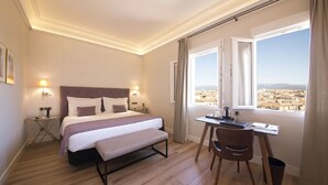 Habitación de hotel con cama, mesitas de noche, escritorio y una silla. Se aprecia una ventana con vistas a un paisaje urbano.