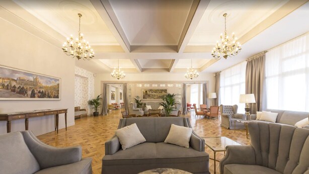 Amplia sala de estar con un sofá gris grande, una mesa de centro y varios sillones. Hay dos candelabros, un cuadro en la pared y un ventanal con cortinas.