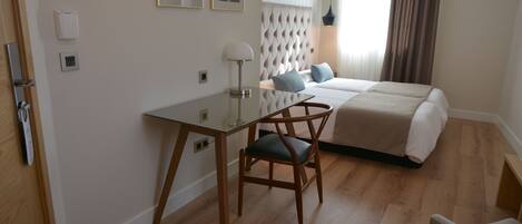 A hotel room with a bed, a desk, and a chair. There are two framed pictures on the wall, a lamp, and a window with curtains.