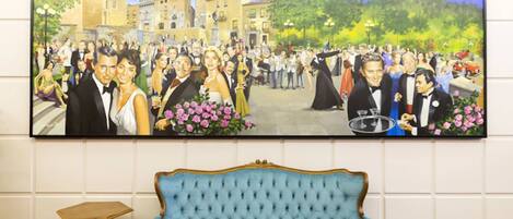 A large painting of a formal event in a square with buildings and a castle in the background, a blue sofa, and a small wooden table in the foreground.