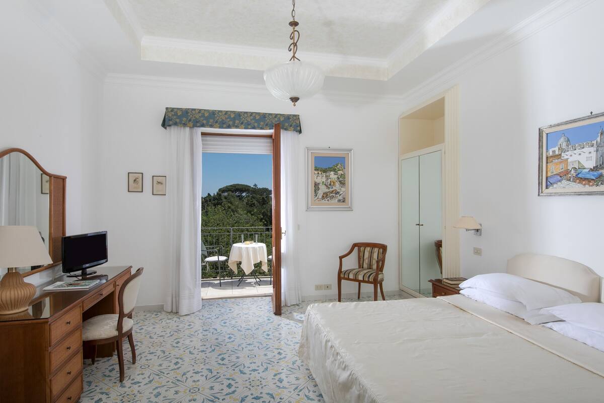 A hotel room with a bed, desk, chair, and a balcony with a table and chairs.