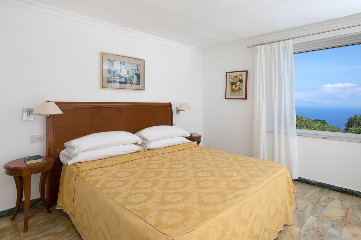 A neatly made bed with a patterned bedspread, a wooden headboard, and two bedside tables with lamps. There are framed pictures on the wall, and a window with a view of greenery and the sky.