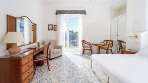A hotel room with a bed, a desk with a lamp, two chairs, a dresser, and a large window with a view of the outdoors. - Hotel Bellavista (Anacapri)