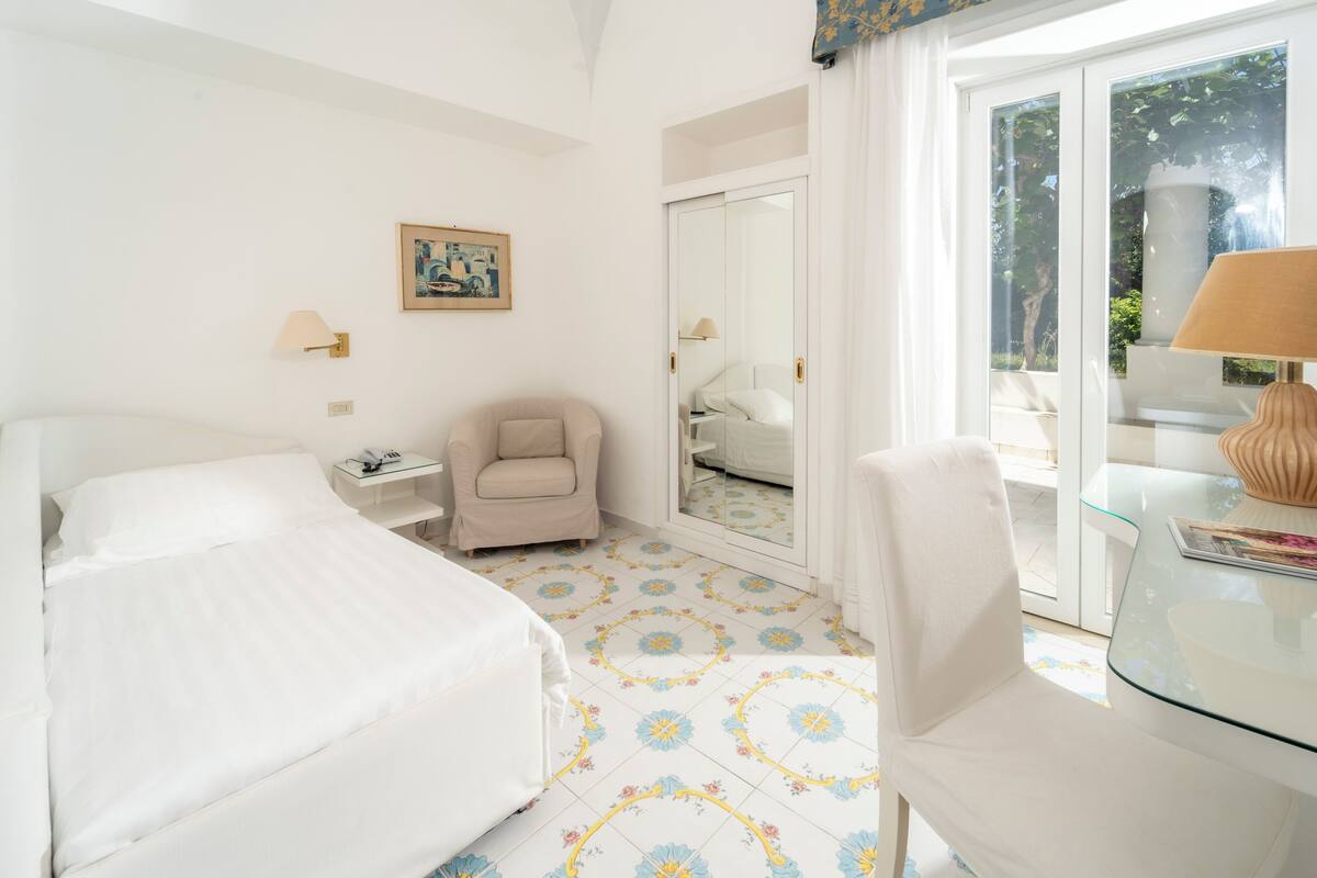 A hotel room with two beds, a chair, a small table, a lamp, a painting, and a glass door leading to a balcony.