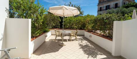 A patio with a table and chairs under an umbrella, surrounded by greenery and a building in the background.