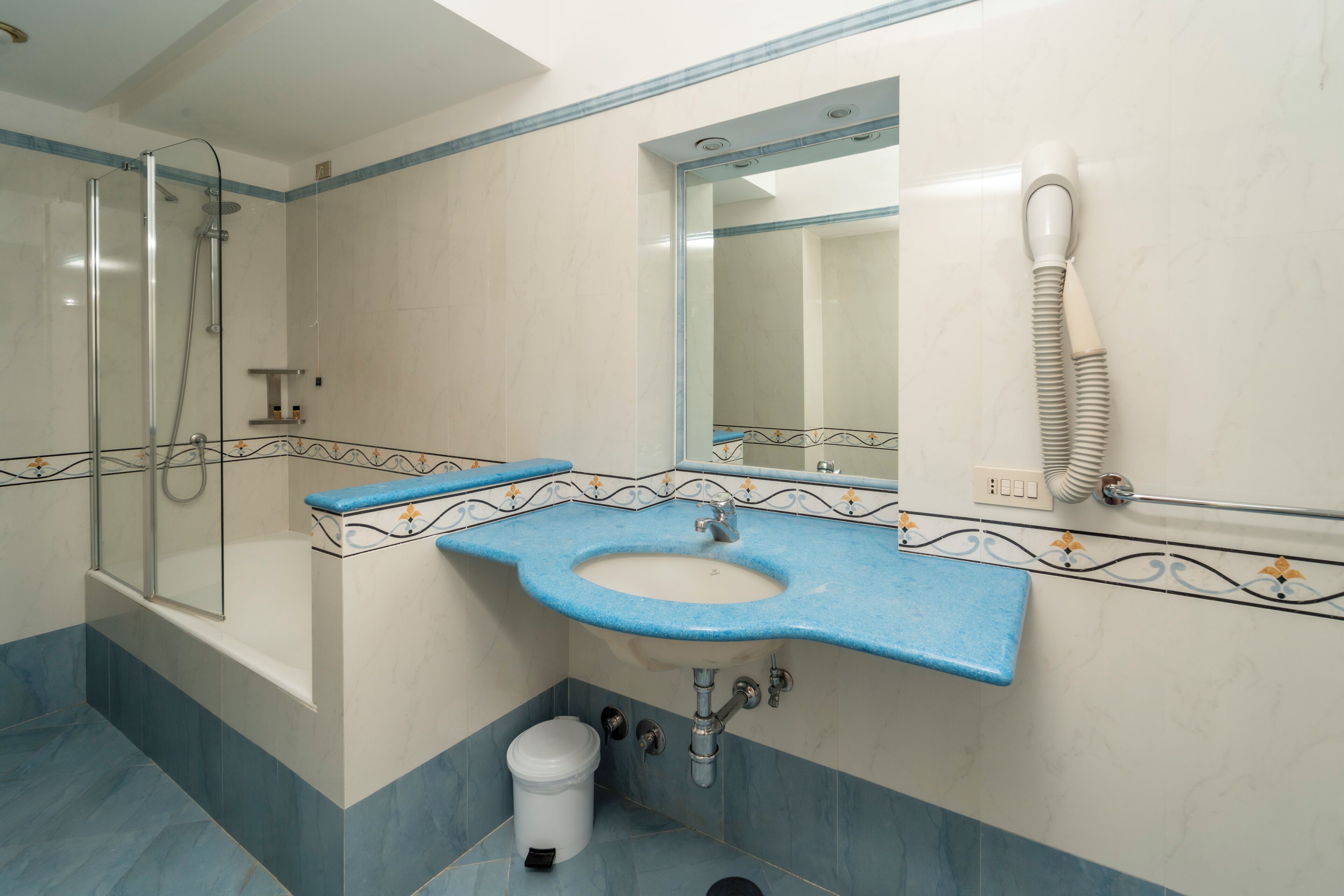 Ein Badezimmer mit blau gefliestem Boden, einem weißen Waschbecken mit blauer Arbeitsplatte, einer Duschkabine aus Glas, einer weißen Toilette und einem Spiegel mit Telefon.