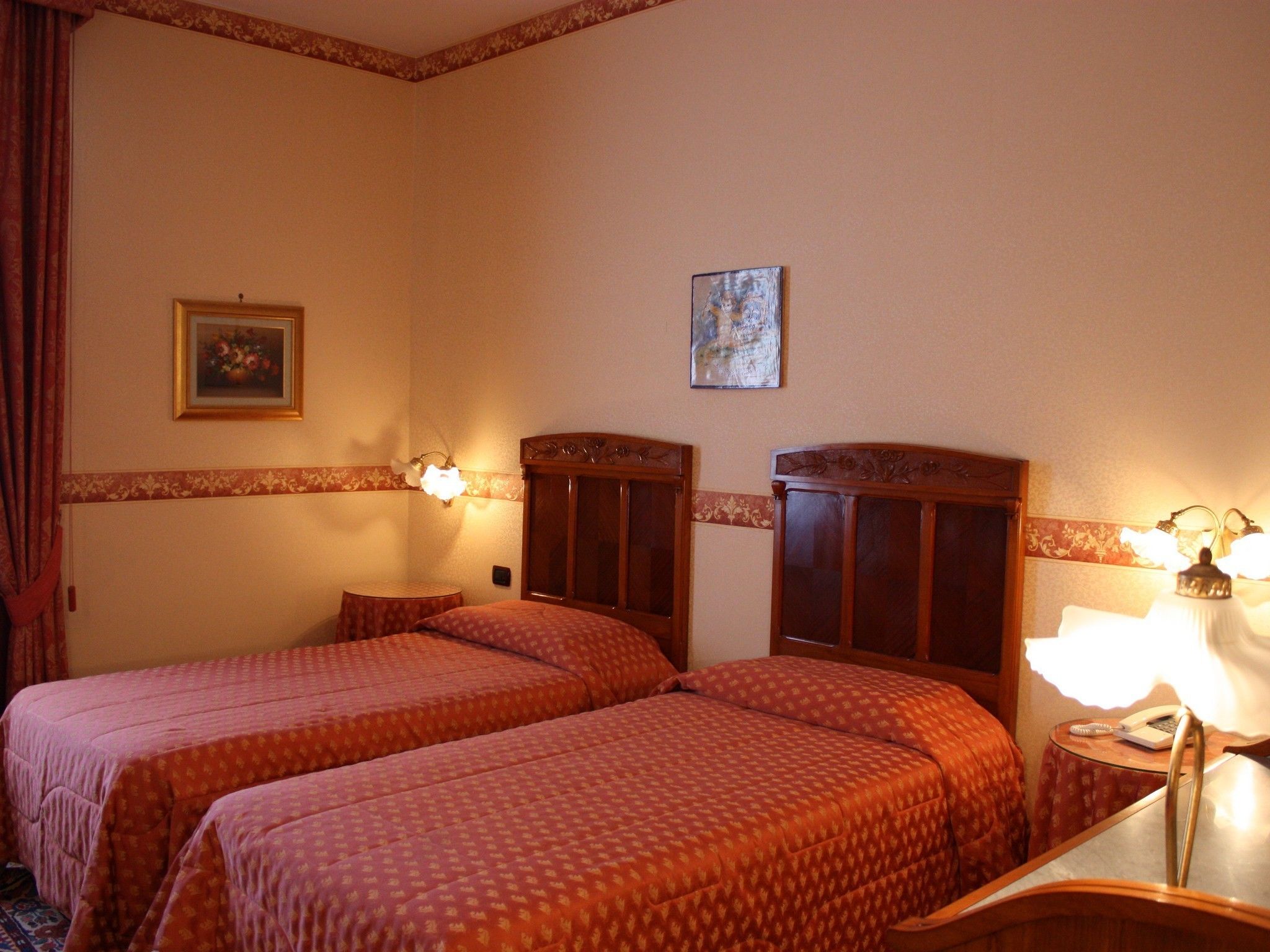 A hotel room with two single beds, a small table with a lamp, a painting on the wall, and a decorative border along the ceiling.