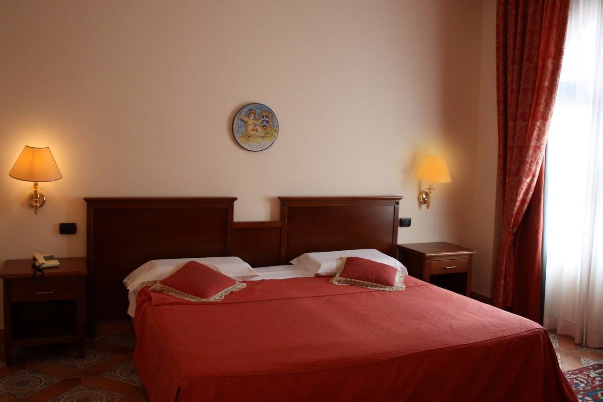 A hotel room with a large bed, two bedside tables, a nightstand, a wall clock, and a window with curtains.