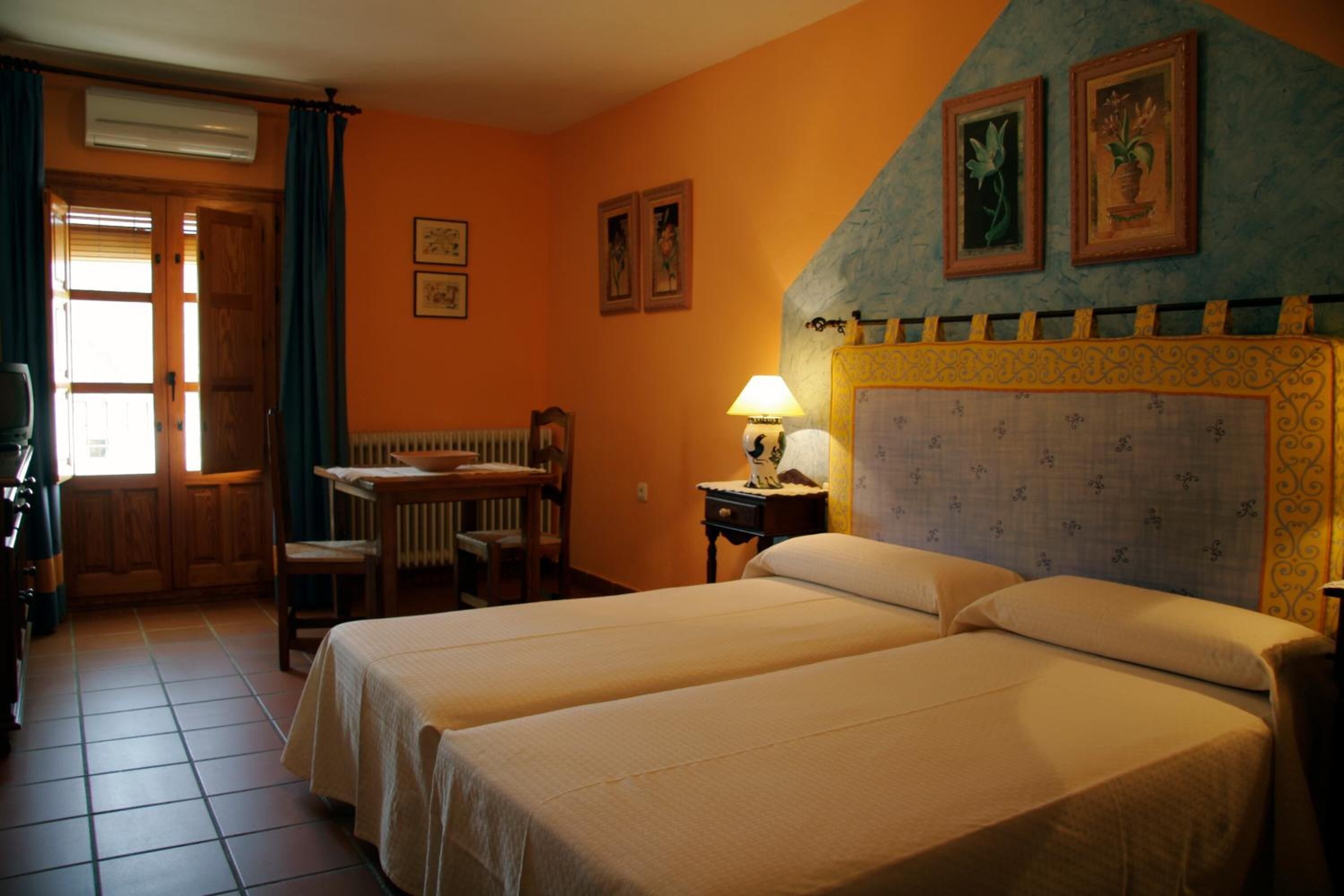 Habitación de hotel con dos camas, un escritorio de madera, una silla, un radiador y una ventana con cortinas.