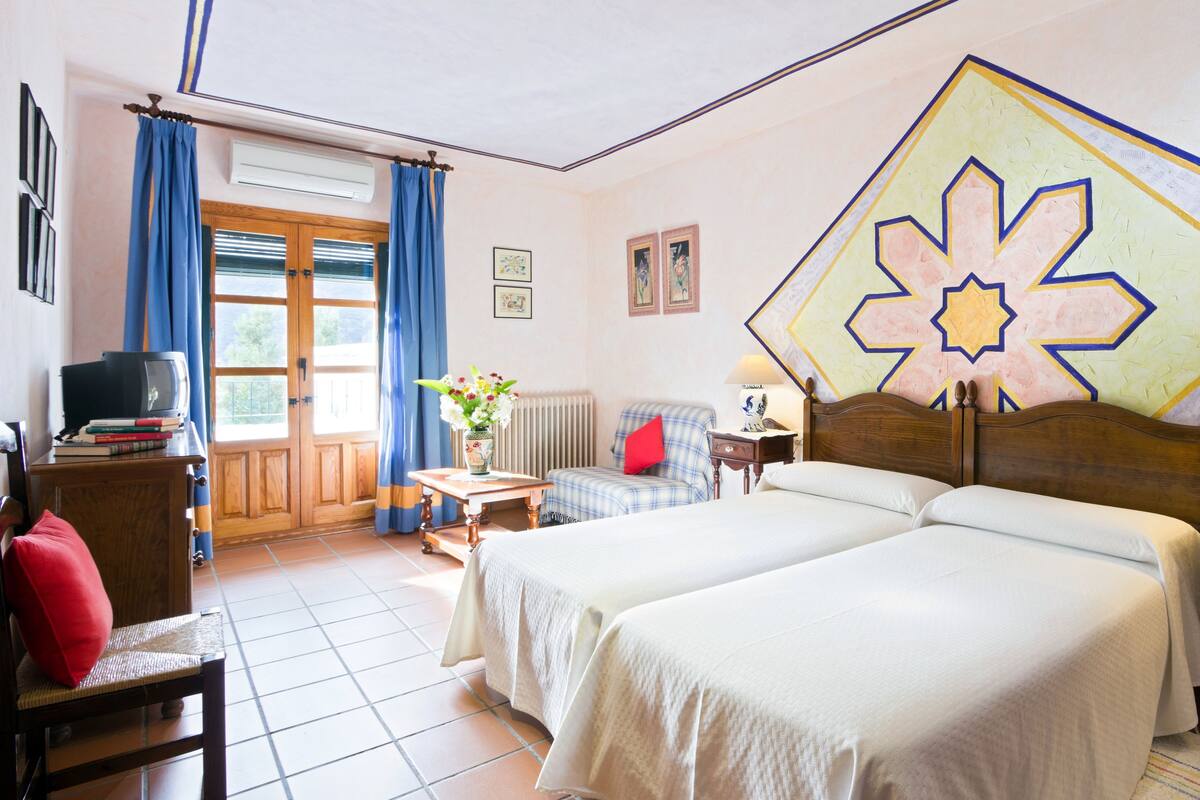 Habitación de hotel con dos camas, televisión, una silla, una mesa con un jarrón de flores y una ventana con cortinas.