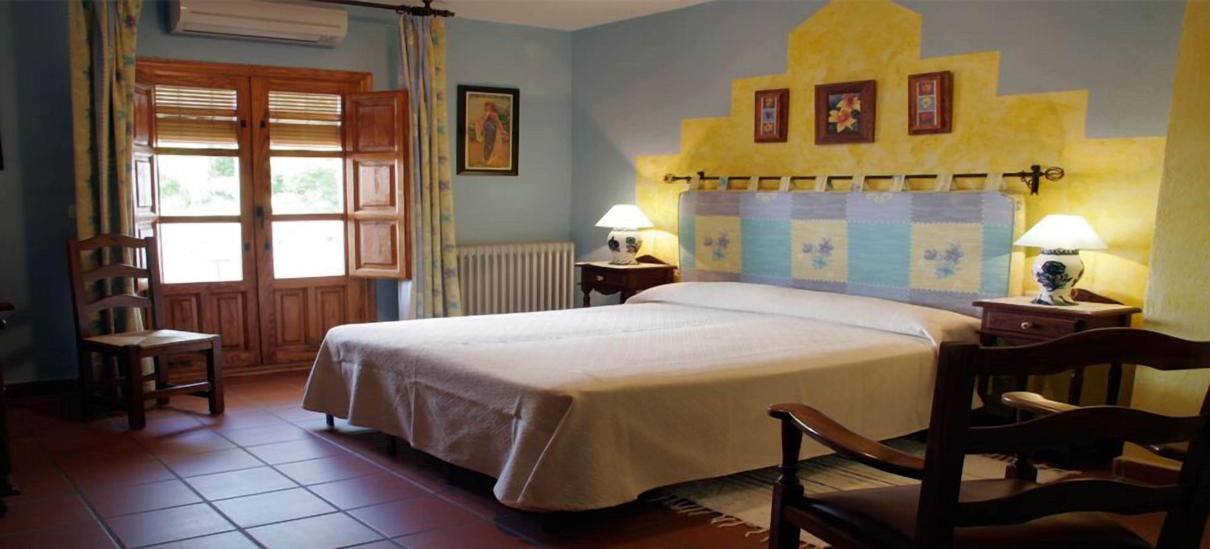 Un dormitorio con una cama, dos mesitas de noche con lámparas, una silla, una ventana con cortinas y un radiador.