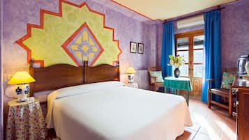 Habitación de hotel con una cama grande, cabecera de madera, dos lámparas de noche, una mesa cubierta con mantel floral, una silla, una mesita con un florero y un televisor. Se observa también una ventana con cortinas azules.