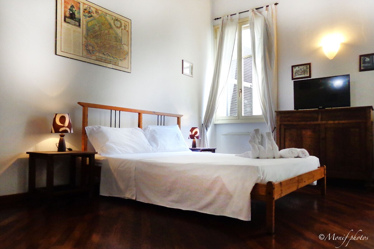 Una cama bien hecha en una habitación con cabecera de madera, una mesita de noche con lámpara, un televisor sobre un mueble de madera y un mapa en la pared.