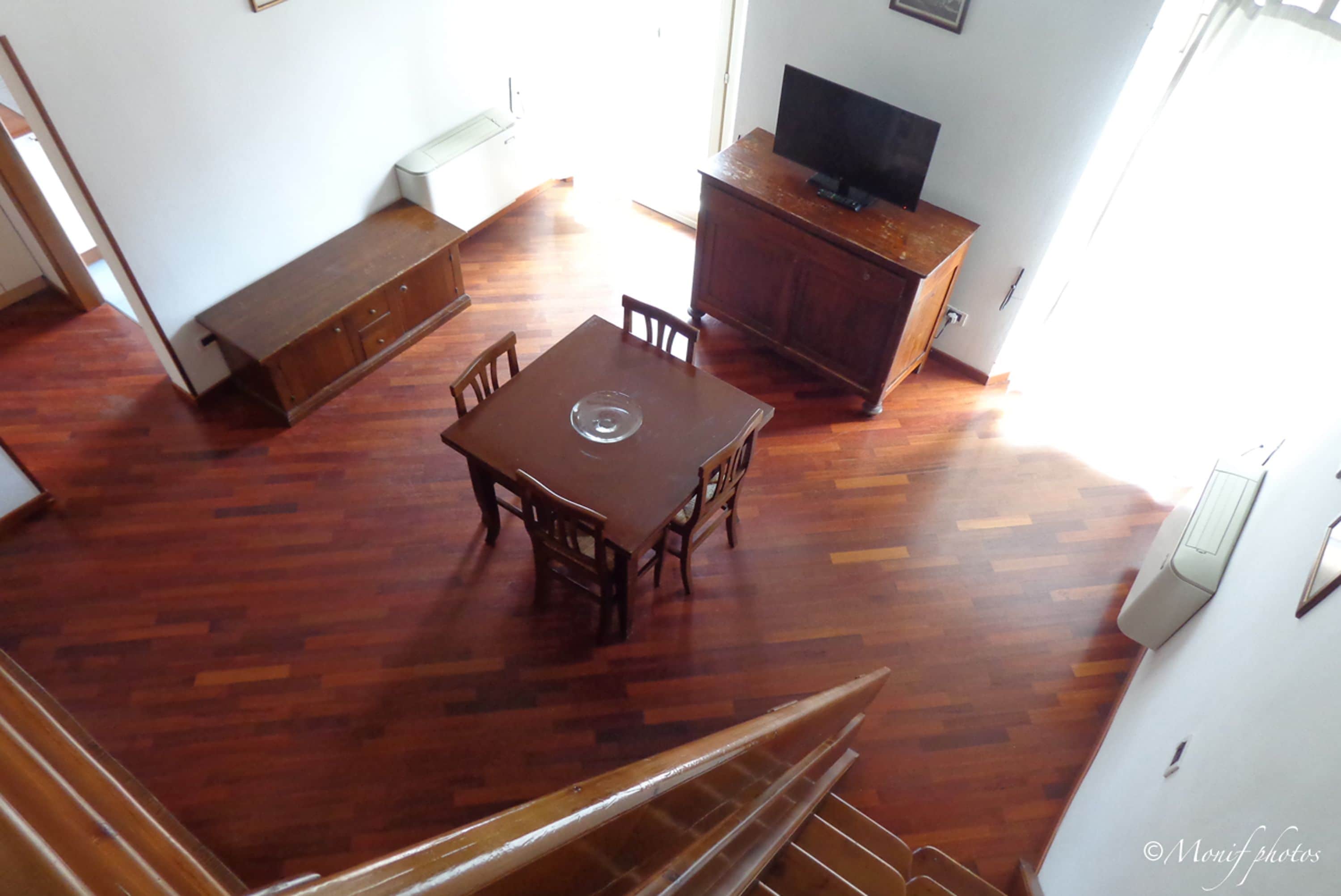 Une pièce spacieuse comprenant une table à manger en bois, des chaises, un banc, un téléviseur posé sur un meuble en bois et un escalier menant à un autre niveau.