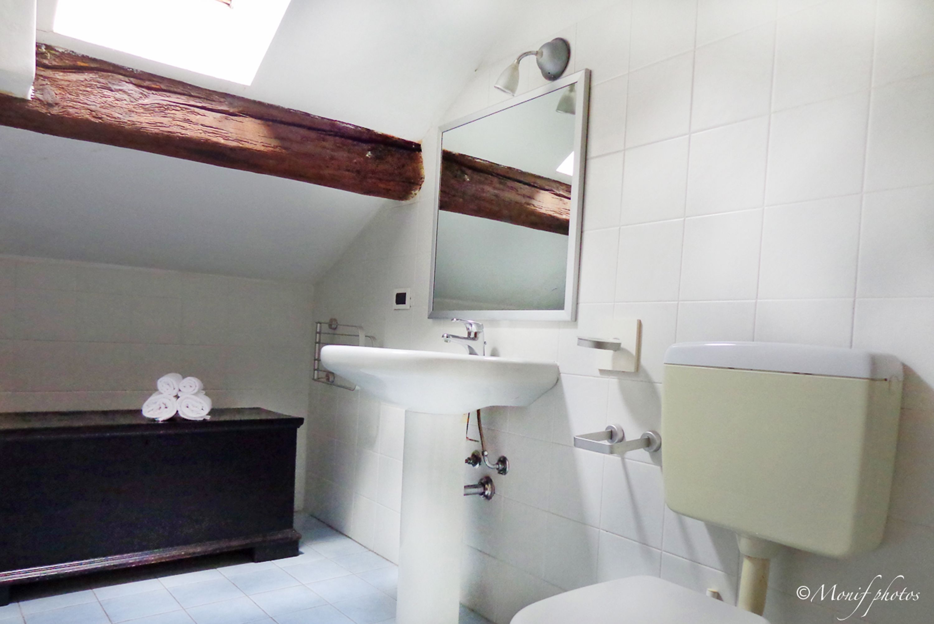 Ein Badezimmer mit weißem Waschbecken, einem Spiegel, einer Toilette und einer dunklen Holzbank.