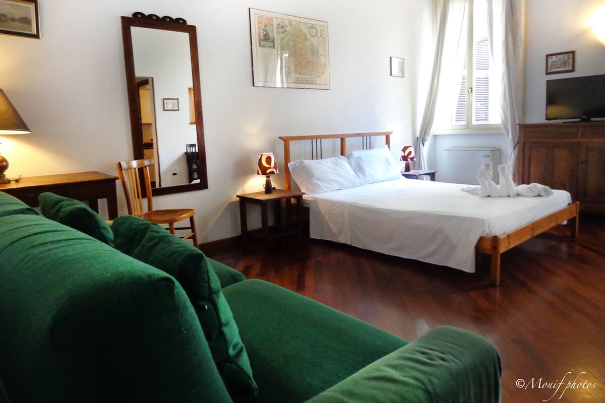 A hotel room with a bed, a green sofa, a wooden nightstand, a lamp, a mirror, and a TV.