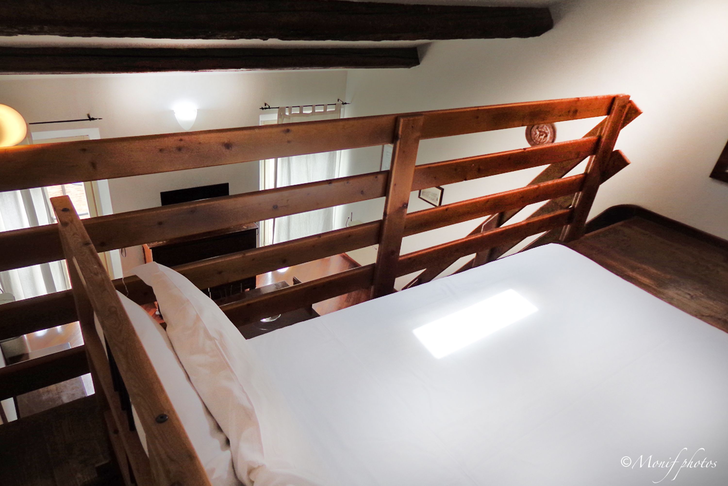 Un cadre de lit en bois avec un matelas blanc, dans une pièce dont on peut voir les poutres apparentes et qui possède un mur blanc.