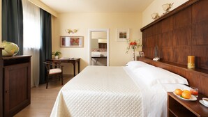 Une chambre d’hôtel avec un grand lit, un bureau avec une chaise, une armoire en bois et une petite table avec des fruits et une boisson.