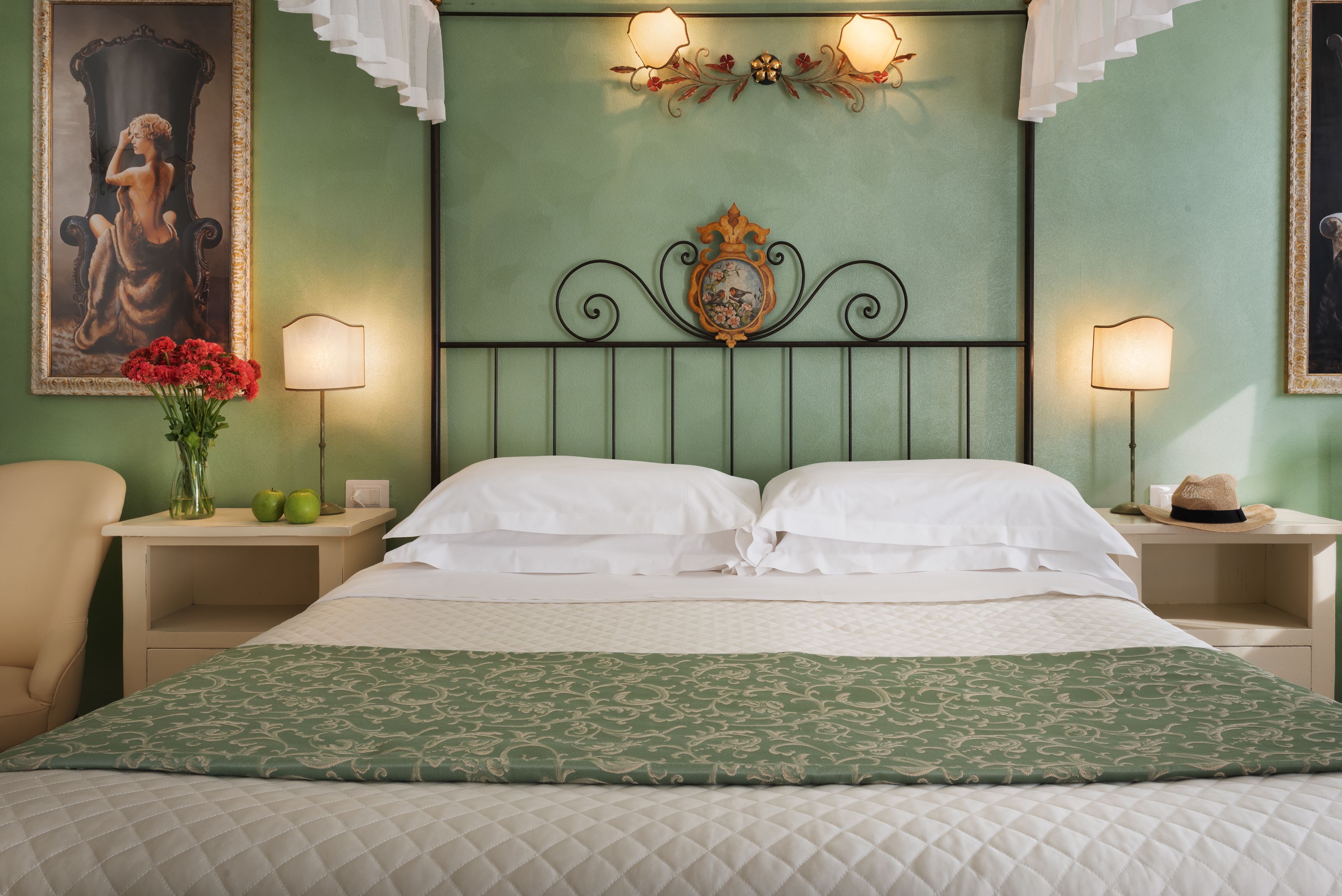 A neatly made bed with white linens and a green patterned throw, flanked by two bedside tables with lamps and a vase of flowers.