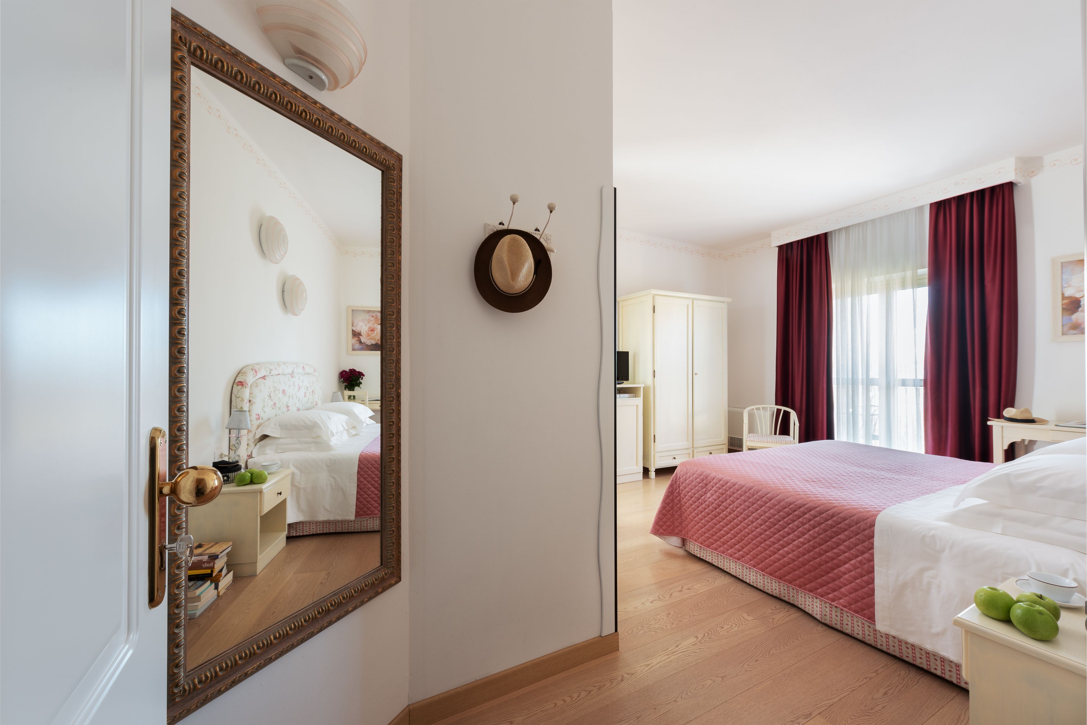 A hotel room with a large mirror, a bed with a red quilt, a nightstand with books, and a wardrobe.